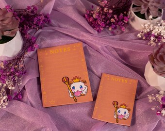 May include: Two orange notepads with the word "NOTES" at the top, featuring a cartoon character with a crown and scepter. The notepads are on a purple fabric with decorative plants.