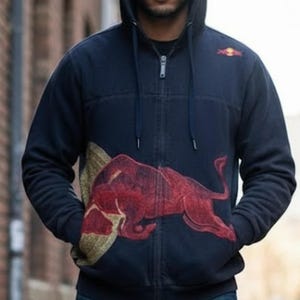 Puede incluir: Sudadera con capucha azul marino con un gran logotipo de toro rojo. La sudadera tiene capucha y bolsillos, y el logotipo de Red Bull también está en la parte superior izquierda del pecho. La persona lleva vaqueros azules.