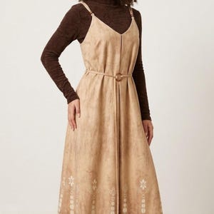 May include: A tan midi dress with a V-neck and thin straps, worn over a brown long-sleeved top. The dress has a decorative belt and a patterned hemline. The model is wearing black trainers and white socks.