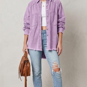 May include: A woman wearing a brown hat, a light purple button-down shirt with fringe detailing, a white top, light blue ripped jeans, and brown boots. She is also carrying a brown leather bag with a shoulder strap.