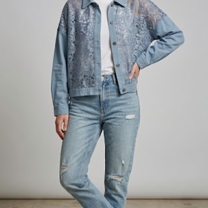 Könnte beinhalten: Hellblaue Jeansjacke mit aufwendigen floralen Spitzendetails auf dem oberen Rücken und den Ärmeln. Die Jacke wird mit hellblauen, zerrissenen Jeans und weißen Turnschuhen kombiniert. Das Outfit ist lässig und stilvoll.