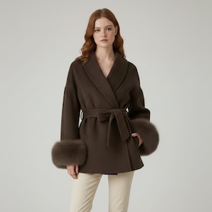 May include: A dark brown wrap coat with a shawl collar and a matching belt. The coat features fur cuffs and is worn over cream-colored pants. The coat is a stylish outerwear garment.
