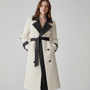 May include: Cream-coloured trench coat with black faux leather accents. The coat features a notched collar, a matching belt, and black trim along the sleeves and front. It has a double-breasted design with black buttons.