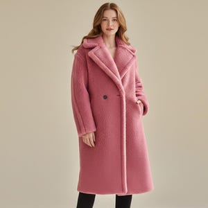 Könnte beinhalten: Ein langer, staubrosafarbener Mantel mit einer strukturierten, weichen Außenseite. Der Mantel hat einen klassischen Reverskragen, einen doppelreihigen Knopfverschluss und zwei Vordertaschen. Der Mantel ist knielang und wirkt warm und bequem.