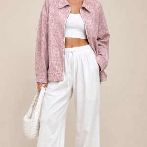 Può includere: Una giacca con zip color rosa polvere con un sottile motivo leopardato, indossata sopra un top corto bianco. L'outfit include pantaloni bianchi a gamba larga a vita alta con coulisse, scarpe da ginnastica bianche e una borsa bianca intrecciata.