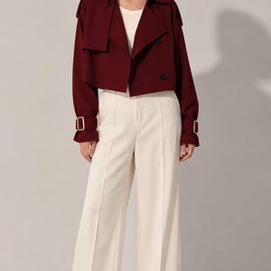 May include: A cropped burgundy trench jacket with a wide collar, double-breasted button closure, and buckled sleeve straps, paired with high-waisted cream-colored wide-leg trousers featuring a center crease and a matching cream-colored top.