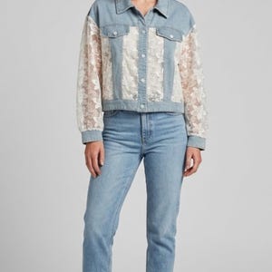 Puede incluir: Chaqueta vaquera azul claro con paneles y mangas de encaje blanco. La chaqueta tiene cuello y cierre de botones. La modelo lleva vaqueros azul claro y zapatillas blancas. El look general es casual y elegante.