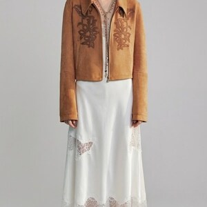 May include: A woman in a tan suede jacket with floral embroidery and a white dress with lace trim. The jacket has a collared neckline and a zip. The dress is a midi length.
