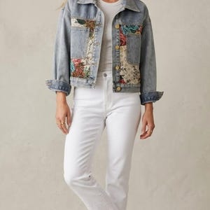 May include: Light blue denim jacket with floral patchwork on the front. The jacket has a classic collar, button closures, and cuffed sleeves. Paired with white jeans and sneakers, creating a casual, stylish outfit.