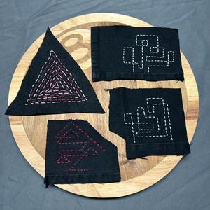 May include: Four black fabric squares with white and red embroidery designs. The designs are geometric shapes, including a triangle, a square, and a rectangle. The squares are arranged on a wooden surface.