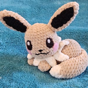 Puede incluir: Un peluche hecho a mano de un personaje de dibujos animados, con un cuerpo marrón claro, grandes orejas con puntas negras y un volante blanco y crema. El juguete tiene mejillas rosadas y ojos negros grandes. El peluche está sobre una superficie azul.
