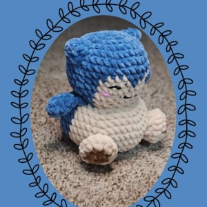 May include: A handmade crochet Snorlax plushie in blue, cream, and brown. The plushie is part of the "Chibi Series" and features a detailed design with closed eyes. The image includes text that reads "Eeveelutionary Crochet" and "Snorlax".