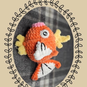 Puede incluir: Un peluche de Magikarp tejido a ganchillo en naranja y blanco, con aletas amarillas y una cresta rosa, de la serie Chibi. El peluche está dentro de un marco ovalado con un diseño de hojas. El texto incluye "Eeveelutionary Crochet" y "Magikarp".