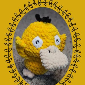 May include: A yellow crocheted plush toy resembling the character Psyduck, featuring a large beige bill, white eyes with black pupils, and a small black tuft of hair on top. The text Chibi Series: Psyduck is displayed above the plush figure.