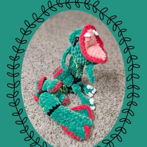 Puede incluir: Un peluche de ganchillo verde, rojo y negro que se asemeja al personaje Rayquaza, con la boca abierta, dientes blancos, detalles amarillos y un acabado de hilo texturizado. La imagen incluye el texto Chibi Series: Rayquaza y Eevolutionary Crochet.