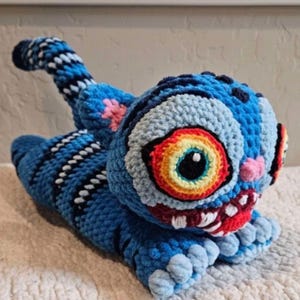 Puede incluir: Una criatura azul de ganchillo hecha a mano con ojos grandes y expresivos y una amplia sonrisa con dientes. El texto "Derpy" es visible, junto con la frase "From Kpop Demon Hunters". El animal de ganchillo tiene una cola rayada y está tumbado.