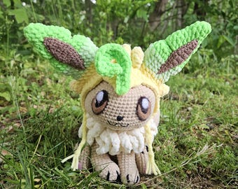 May include: A handmade crochet plush toy of a Leafeon Pokémon character. The plush features green, brown, and yellow yarn, with large eyes and leaf-like ears. The toy is sitting in a grassy area, showcasing its detailed craftsmanship.