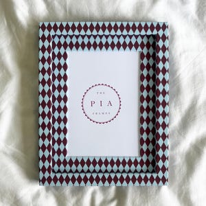May include: Rectangular picture frame with a repeating diamond pattern in burgundy and light blue. The frame has a white interior with the text "THE PIA FRAMES" in a circular design.