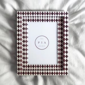 Könnte beinhalten: Rechteckiger Bilderrahmen mit einem sich wiederholenden Rautenmuster in Burgunderrot und Weiß. Der Rahmen hat einen weißen Innenraum mit dem Text "THE PIA FRAMES" in einem kreisförmigen Design.
