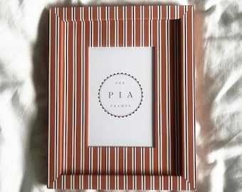 Könnte beinhalten: Ein rechteckiger Bilderrahmen mit vertikalem Streifenmuster in Terrakotta, Weiß und Schwarz. In der Mitte des Rahmens befindet sich ein weißer Einleger mit einem kreisförmigen Logo, das den Text THE PIA FRAMES trägt.