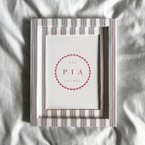 Puede incluir: Marco de fotos rectangular con un patrón de rayas rojas, blancas y azules. El marco tiene un interior blanco con el texto "THE PIA FRAMES" en un diseño circular.