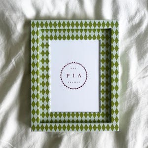 Könnte beinhalten: Ein rechteckiger Bilderrahmen mit einem grün-weißen Rautenmuster. Der Rahmen hat einen weißen Innenraum mit dem Text "THE PIA FRAMES" in einem kreisförmigen Design.