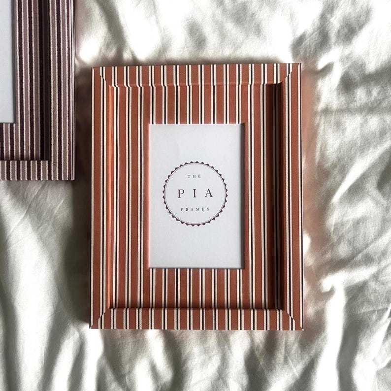May include: A rectangular picture frame with a striped pattern in shades of brown, white, and red. The frame has a white interior with the text "THE PIA FRAMES" in a circular design. The frame is designed for displaying photos or artwork.