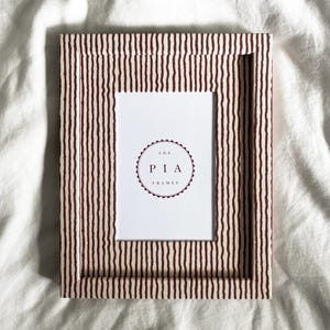 Puede incluir: Un marco de fotos rectangular con un patrón de rayas en tonos marrón y crema. El marco tiene un inserto blanco con el texto "THE PIA FRAMES" en un diseño circular. El marco está diseñado para contener una fotografía.