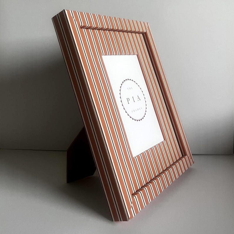 May include: A rectangular picture frame with a striped pattern in shades of brown and white. The frame has a built-in easel stand. The text "THE PIA FRAMES" is visible on the white insert.