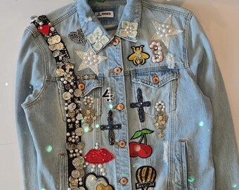 Op de afbeelding: Lichtblauwe spijkerjas versierd met diverse patches, waaronder sterren, kruisen, een bij en een kers. De jas heeft een zwarte riem versierd met munten en parels. Decoratief en modieus.