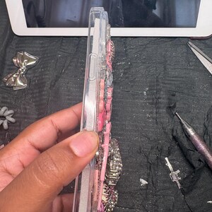 Puede incluir: Una funda de tel&eacute;fono de pl&aacute;stico transparente decorada con una densa disposici&oacute;n de adornos rosas, blancos y plateados. Los elementos decorativos incluyen varias formas, texturas y patrones, como estampado de leopardo y acabados brillantes.