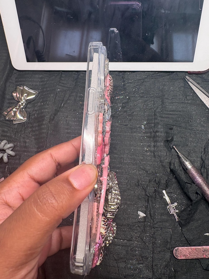 Puede incluir: Una funda de tel&eacute;fono de pl&aacute;stico transparente decorada con una densa disposici&oacute;n de adornos rosas, blancos y plateados. Los elementos decorativos incluyen varias formas, texturas y patrones, como estampado de leopardo y acabados brillantes.