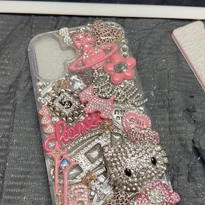 Puede incluir: Una funda de tel&eacute;fono transparente decorada con una densa disposici&oacute;n de dijes rosas y plateados, que incluye una figura de gato cubierta de pedrer&iacute;a, una flor rosa, un dije de frasco de perfume con el n&uacute;mero 5 y varios detalles de leopardo.