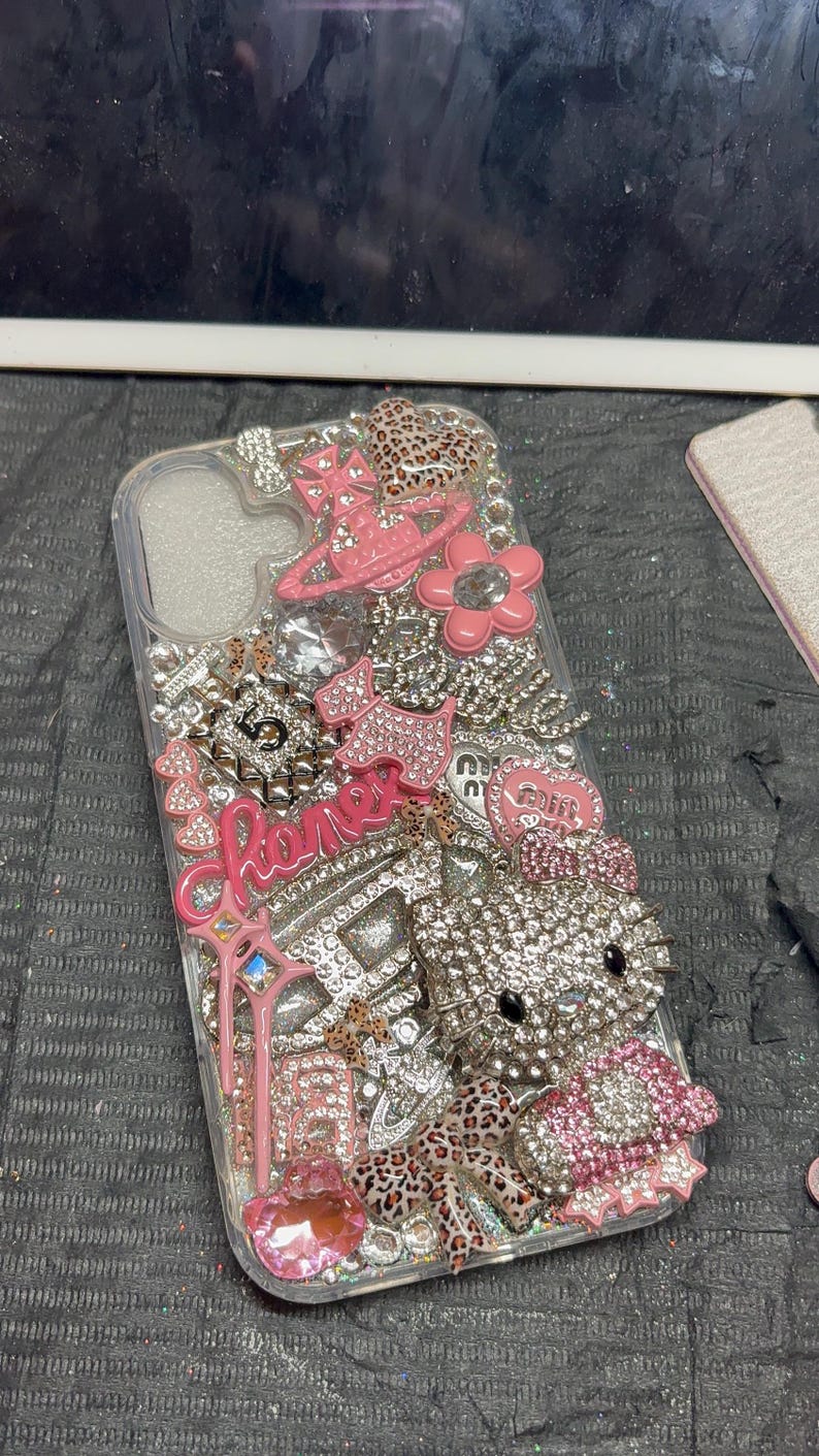 Puede incluir: Una funda de tel&eacute;fono transparente decorada con una densa disposici&oacute;n de dijes rosas y plateados, que incluye una figura de gato cubierta de pedrer&iacute;a, una flor rosa, un dije de frasco de perfume con el n&uacute;mero 5 y varios detalles de leopardo.