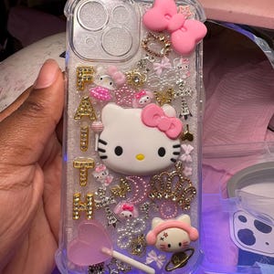 May include: A clear phone case with a Hello Kitty design. The case is decorated with rhinestones, glitter, and other embellishments. The word "FAITH" is spelled out in gold letters on the case.