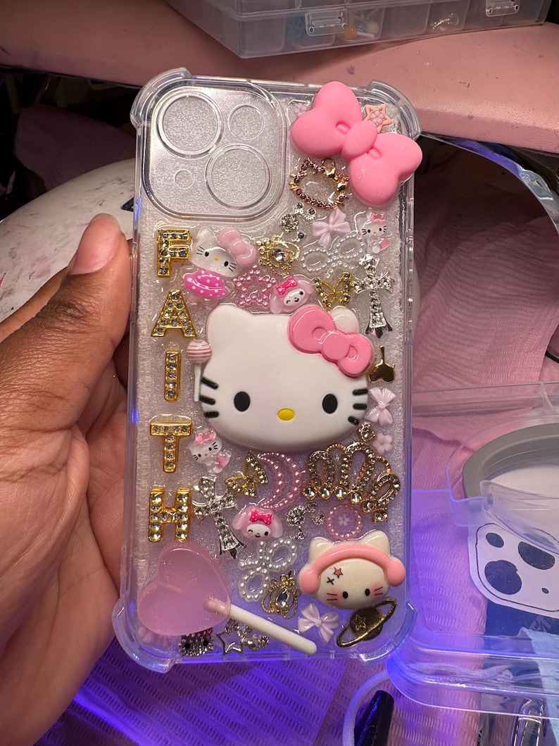 May include: A clear phone case with a Hello Kitty design. The case is decorated with rhinestones, glitter, and other embellishments. The word "FAITH" is spelled out in gold letters on the case.