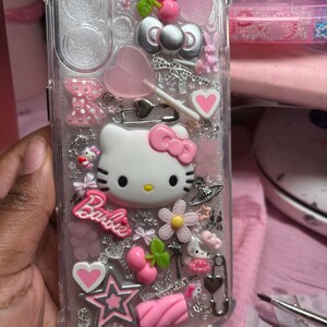 May include: A clear phone case with a Hello Kitty design. The case is decorated with pink, white, and silver glitter, as well as various charms, including a lollipop, a heart, a star, and a cherry. The text "Barbie" is also visible on the case.