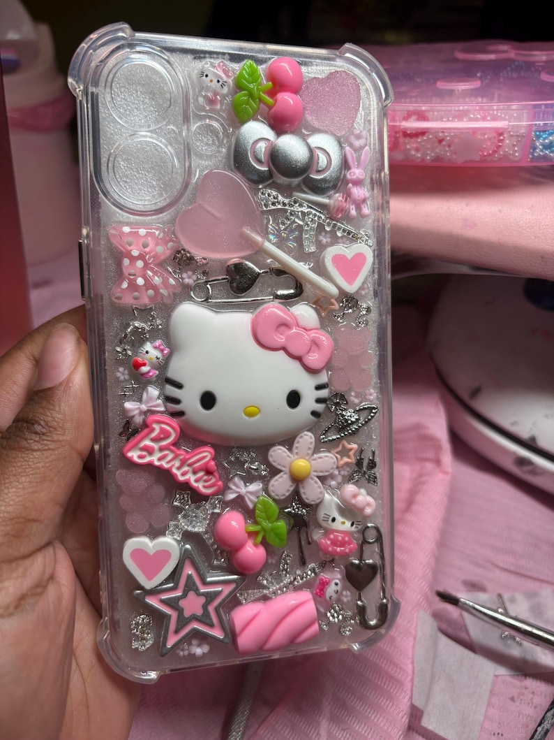 May include: A clear phone case with a Hello Kitty design. The case is decorated with pink, white, and silver glitter, as well as various charms, including a lollipop, a heart, a star, and a cherry. The text "Barbie" is also visible on the case.