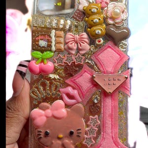 Op de afbeelding: Transparante telefoonhoes versierd met roze Hello Kitty, strikken, sterren en andere bedels. De hoes heeft een glitterachtergrond en heeft een roze kruisontwerp met een klein label. Extra versieringen zijn een chocoladereep en teddybeerfiguren.