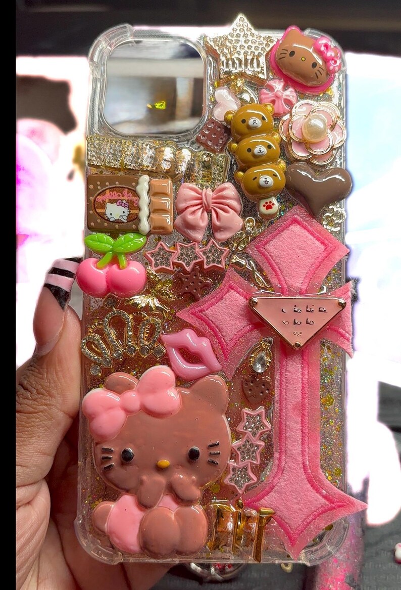 Op de afbeelding: Transparante telefoonhoes versierd met roze Hello Kitty, strikken, sterren en andere bedels. De hoes heeft een glitterachtergrond en heeft een roze kruisontwerp met een klein label. Extra versieringen zijn een chocoladereep en teddybeerfiguren.
