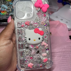 May include: A clear phone case with a Hello Kitty design. The case is decorated with pink, white, and silver glitter, rhinestones, and charms. The case also features a pink bow and a Hello Kitty charm.