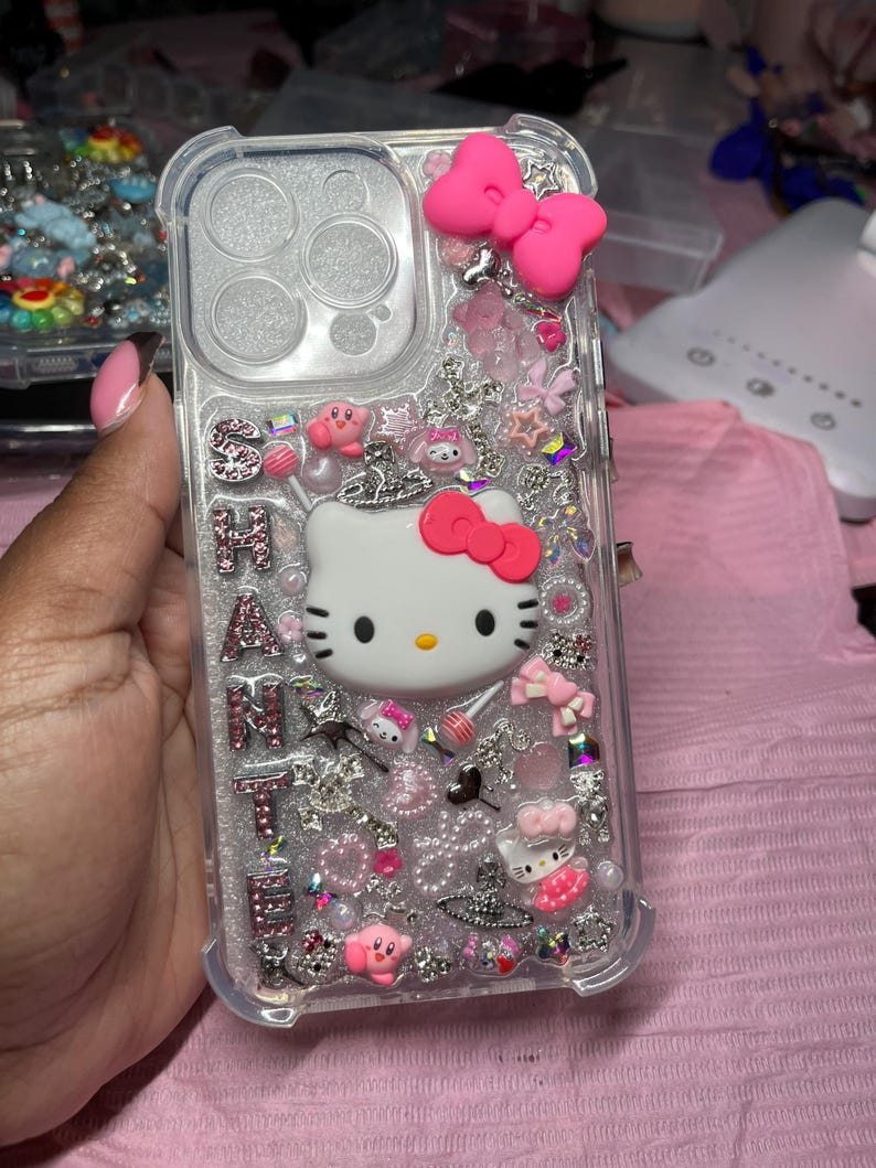 May include: A clear phone case with a Hello Kitty design. The case is decorated with pink, white, and silver glitter, rhinestones, and charms. The case also features a pink bow and a Hello Kitty charm.