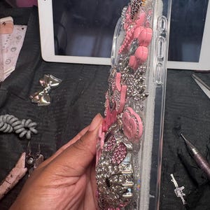 Puede incluir: Una funda de tel&eacute;fono transparente decorada con una densa disposici&oacute;n de pedrer&iacute;a rosa y plateada, peque&ntilde;os dijes rosas en forma de coraz&oacute;n y una figura de gato con cristales rosas y plateados en la parte posterior.