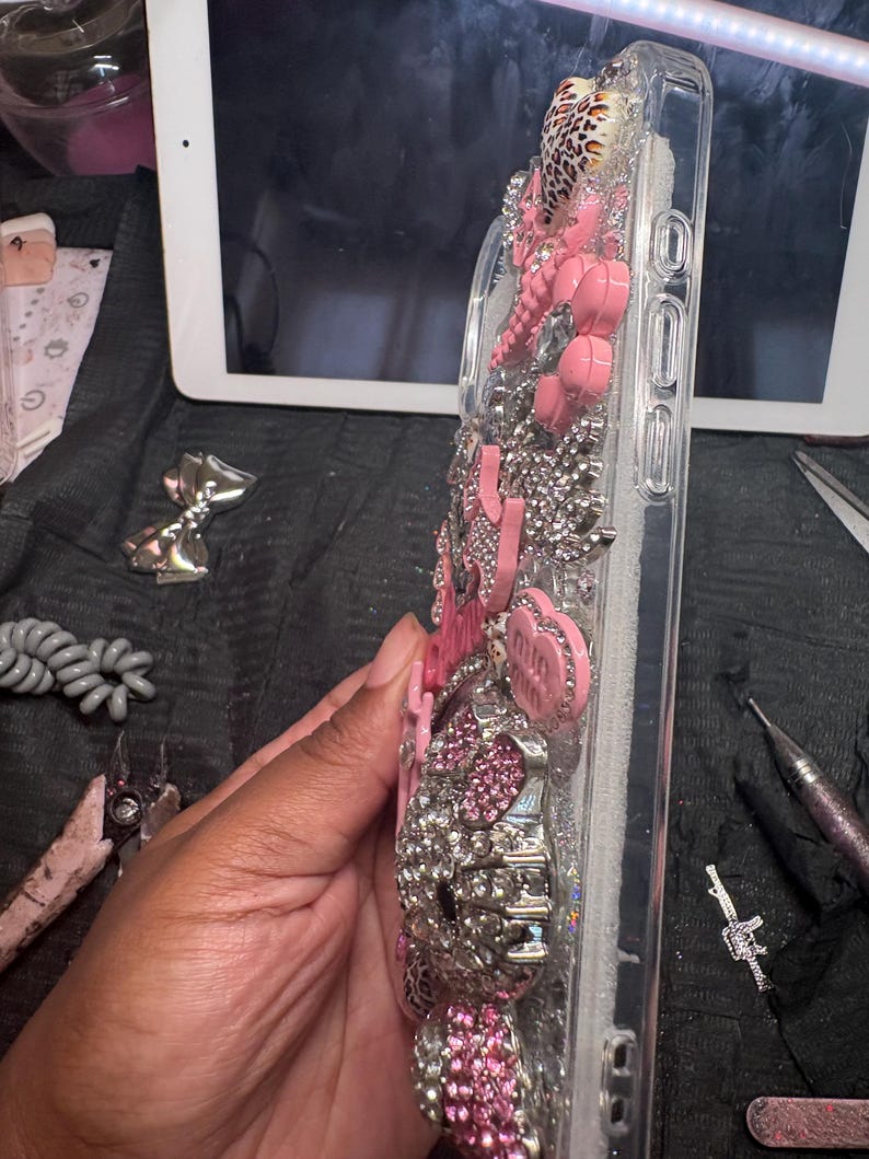 Puede incluir: Una funda de tel&eacute;fono transparente decorada con una densa disposici&oacute;n de pedrer&iacute;a rosa y plateada, peque&ntilde;os dijes rosas en forma de coraz&oacute;n y una figura de gato con cristales rosas y plateados en la parte posterior.