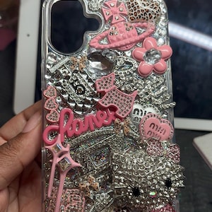 Puede incluir: Una funda de tel&eacute;fono transparente decorada con una densa disposici&oacute;n de pedrer&iacute;a plateada, dijes rosas y varias figuras en miniatura. El dise&ntilde;o incluye un logotipo rosa en cursiva, un personaje de gato cubierto de cristales y detalles de estampado de leopardo.