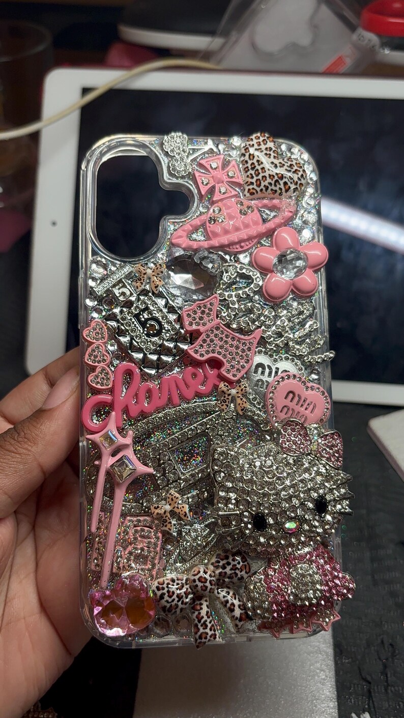 Puede incluir: Una funda de tel&eacute;fono transparente decorada con una densa disposici&oacute;n de pedrer&iacute;a plateada, dijes rosas y varias figuras en miniatura. El dise&ntilde;o incluye un logotipo rosa en cursiva, un personaje de gato cubierto de cristales y detalles de estampado de leopardo.
