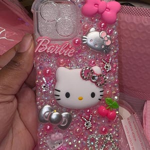 May include: A pink and clear phone case with a Hello Kitty design, rhinestones, and the word "Barbie" on it.
