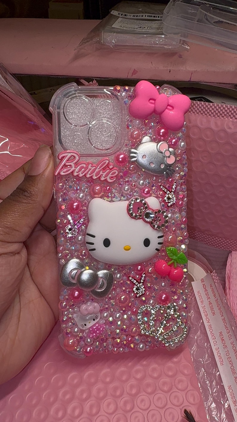 May include: A pink and clear phone case with a Hello Kitty design, rhinestones, and the word "Barbie" on it.