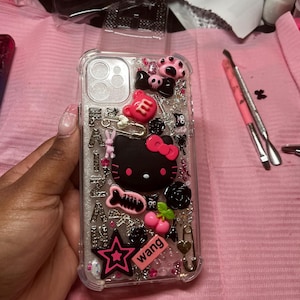 May include: A clear phone case with a glittery background and various pink, black, and white embellishments. The embellishments include a Hello Kitty character, a star, a cherry, a paw print, a safety pin, and a "wang" sticker.