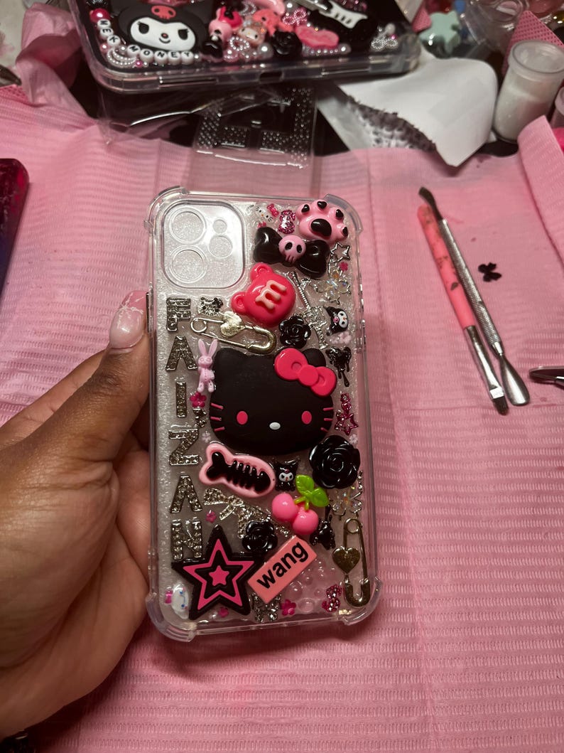 May include: A clear phone case with a glittery background and various pink, black, and white embellishments. The embellishments include a Hello Kitty character, a star, a cherry, a paw print, a safety pin, and a "wang" sticker.